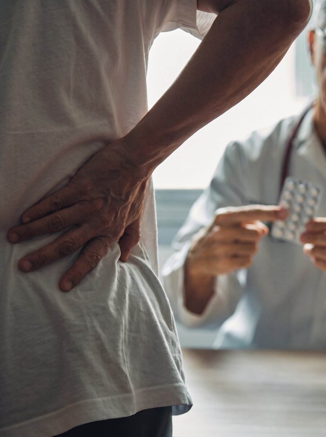 Mujer de espaldas con las manos en las lumbares mientras un médico le ofrece unas pastillas al otro lado del escritorio