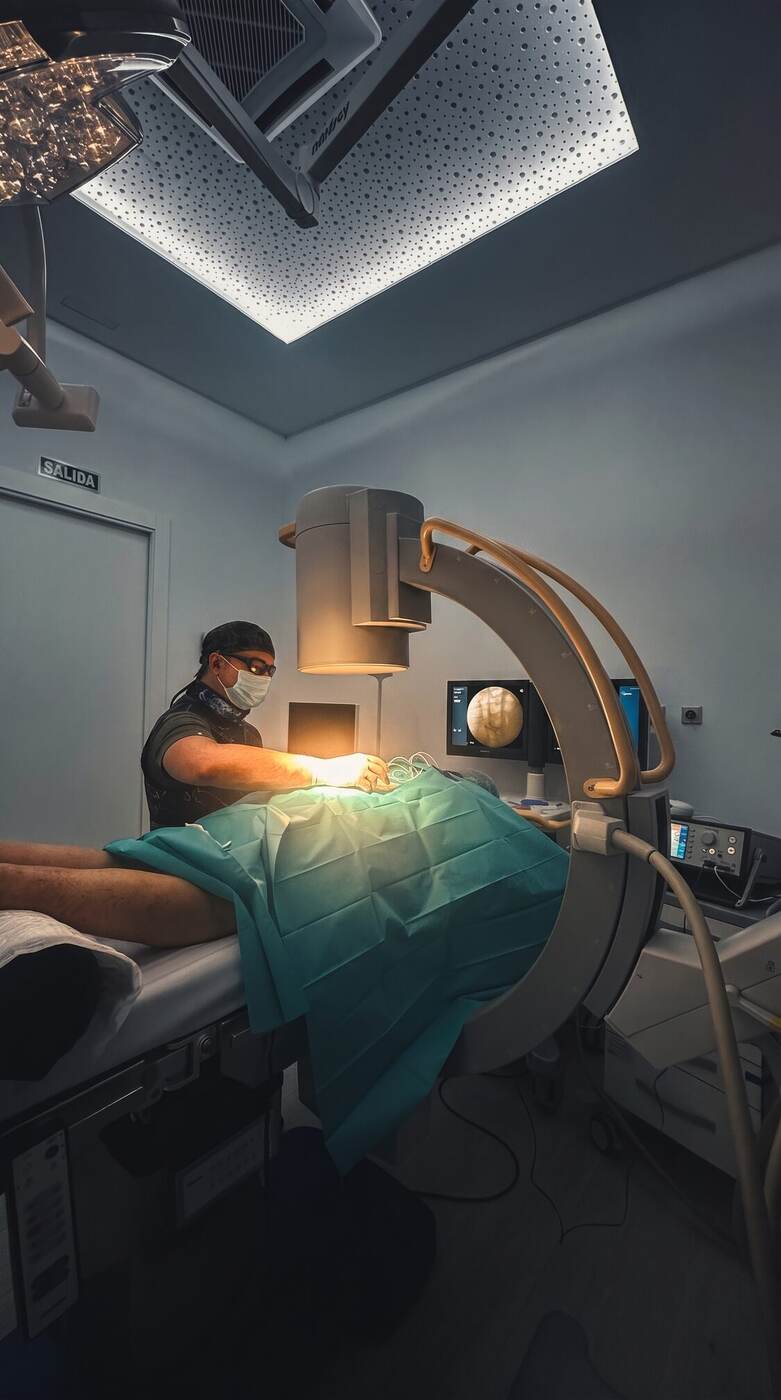 El Dr. Martín Rodríguez Banqueri trabajando bajo el arco de escopia en la sala de intervencionismo de Clínica Dolentia; en los monitores, la imagen radioscópica de la columna lumbar por la que salen las raíces del nervio ciático