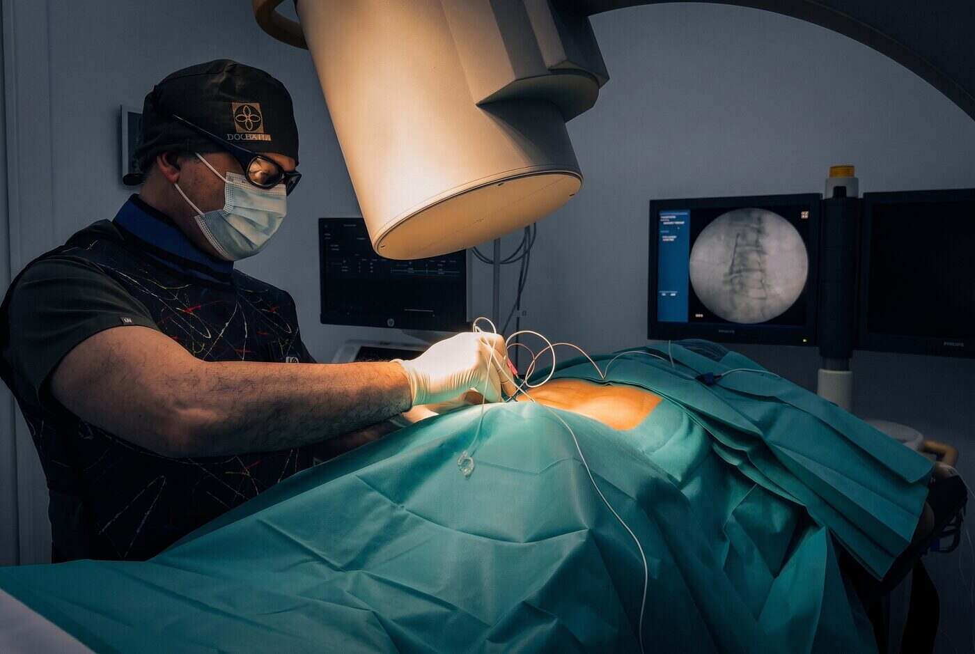 El Dr. Martín Rodríguez Banqueri realizando un procedimiento percutáneo guiado por escopia sobre la columna en la sala de intervencionismo de Clínica Dolentia; en el monitor, la imagen radioscópica que guía la técnica sobre las articulaciones cervicales