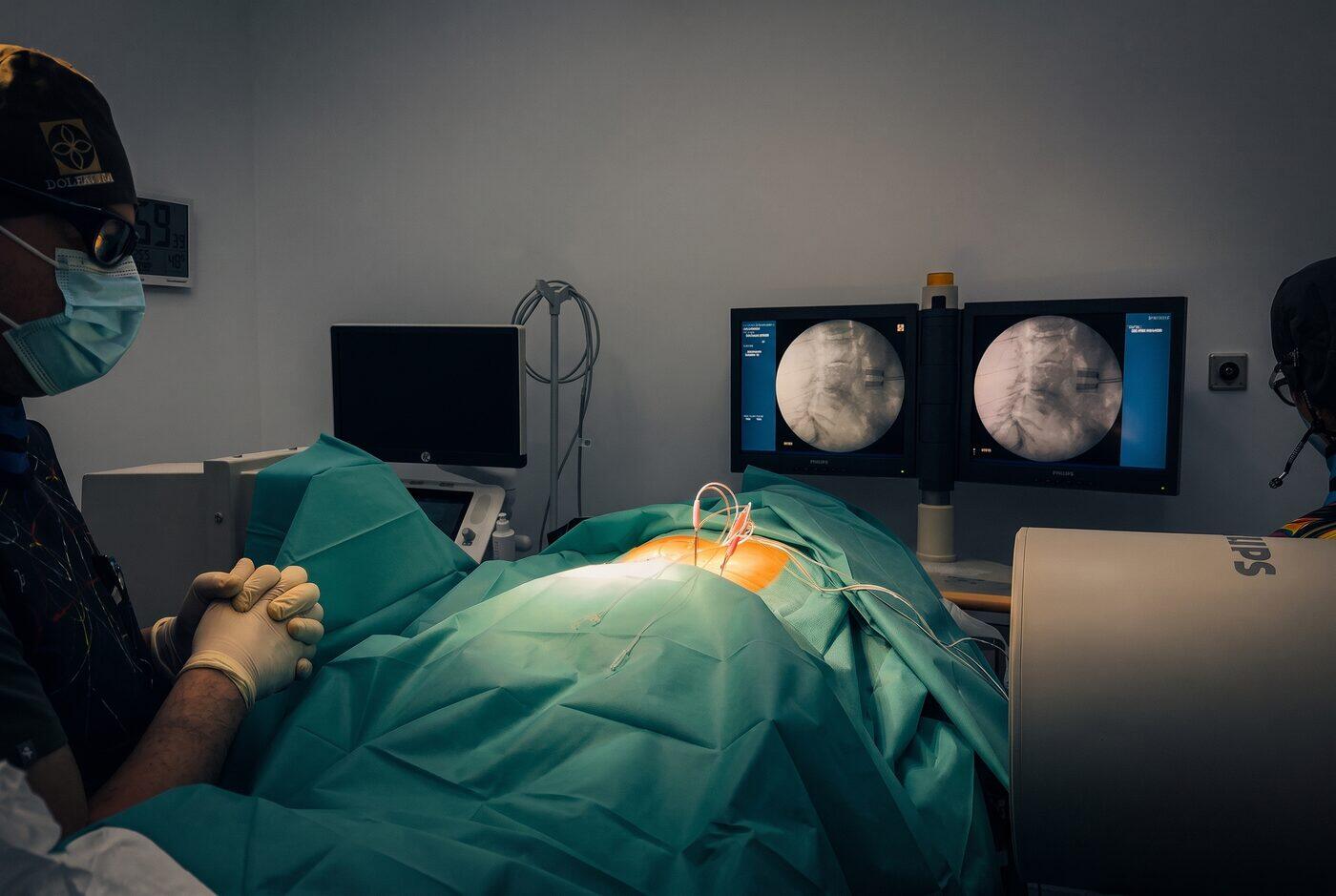 El Dr. Martín Rodríguez Banqueri realizando una técnica percutánea guiada por escopia sobre estructuras nerviosas de la columna en la sala de intervencionismo de Clínica Dolentia; en los dos monitores, la imagen radioscópica que guía el procedimiento
